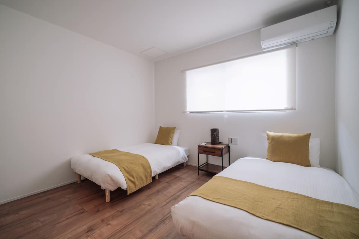 A room with two beds, wooden flooring, white walls, a window, and an air conditioning unit.
