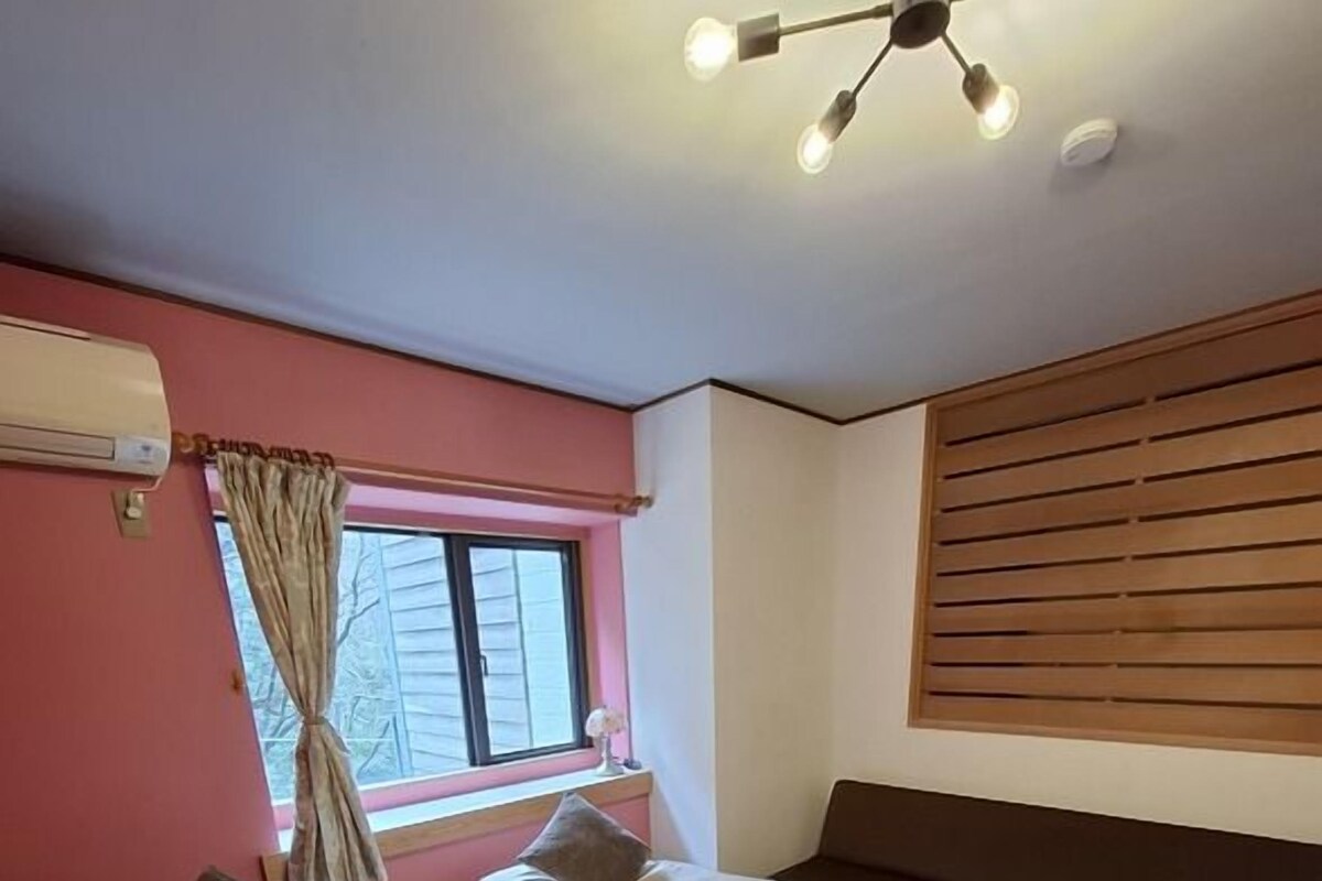 A room with two beds, a window with curtains, a ceiling light fixture, and a wooden floor.