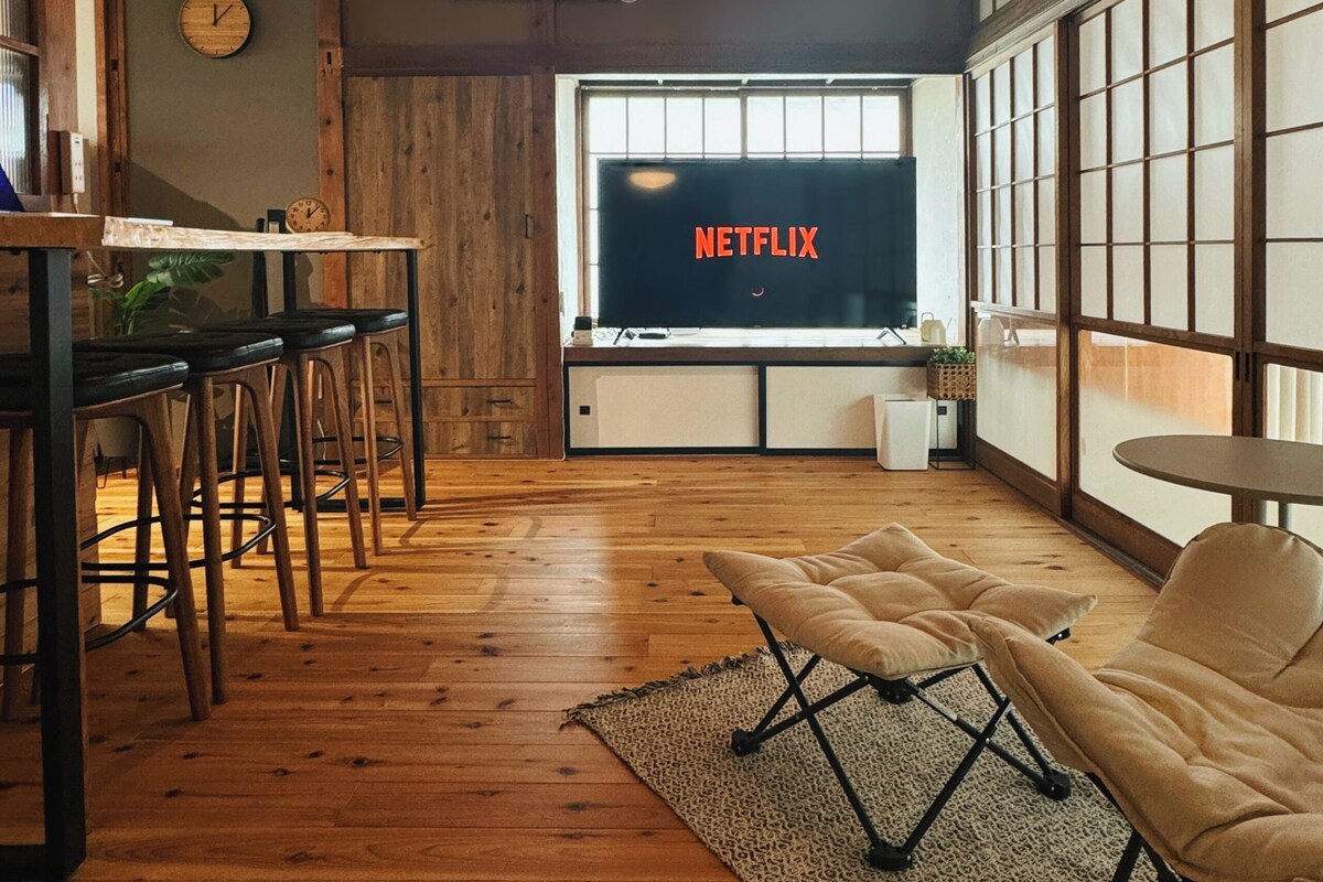 Una habitación con techo de madera, un televisor mostrando 'NETFLIX', una zona de comedor con taburetes y un ventanal grande.