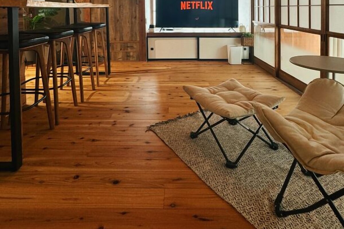 Una habitación con techo de madera, un televisor mostrando 'NETFLIX', una zona de comedor con taburetes y un ventanal grande.
