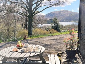 Ein Picknicktisch mit Essen und Tee, ein Hund und ein malerischer Blick auf einen See und Berge.