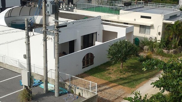 Um edifício de vários andares com telhado plano, uma pequena área de jardim com uma árvore e arbustos, e um estacionamento cercado.