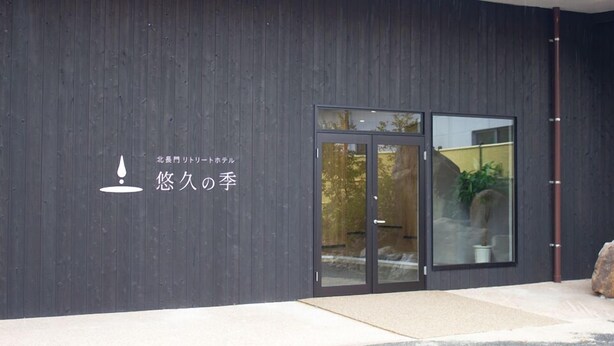Fachada moderna de un edificio con paneles exteriores de madera oscura, puerta de vidrio y una pequeña planta en maceta a la derecha.
