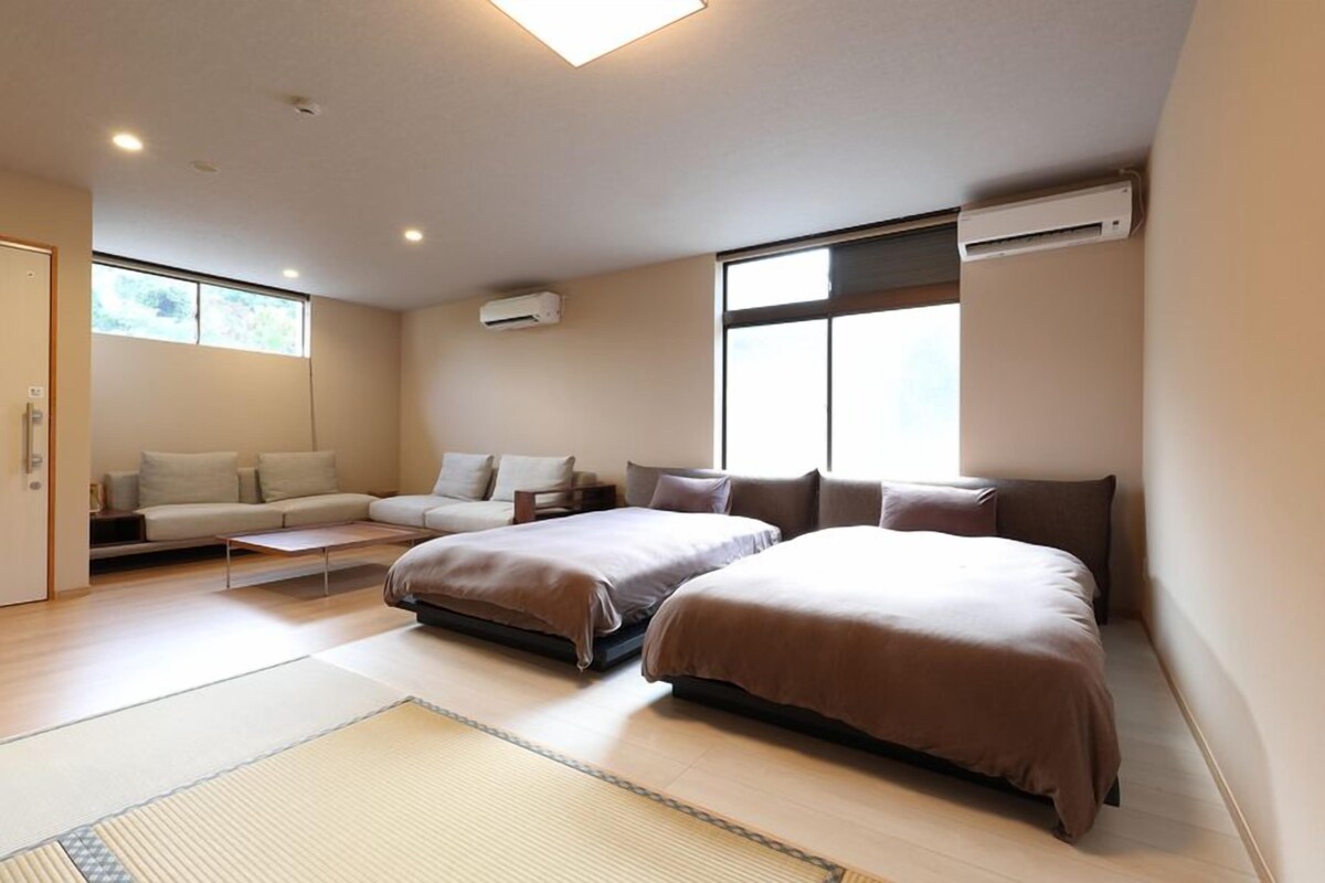 A modern hotel room with two beds, a sofa, a small table, and a large window.
