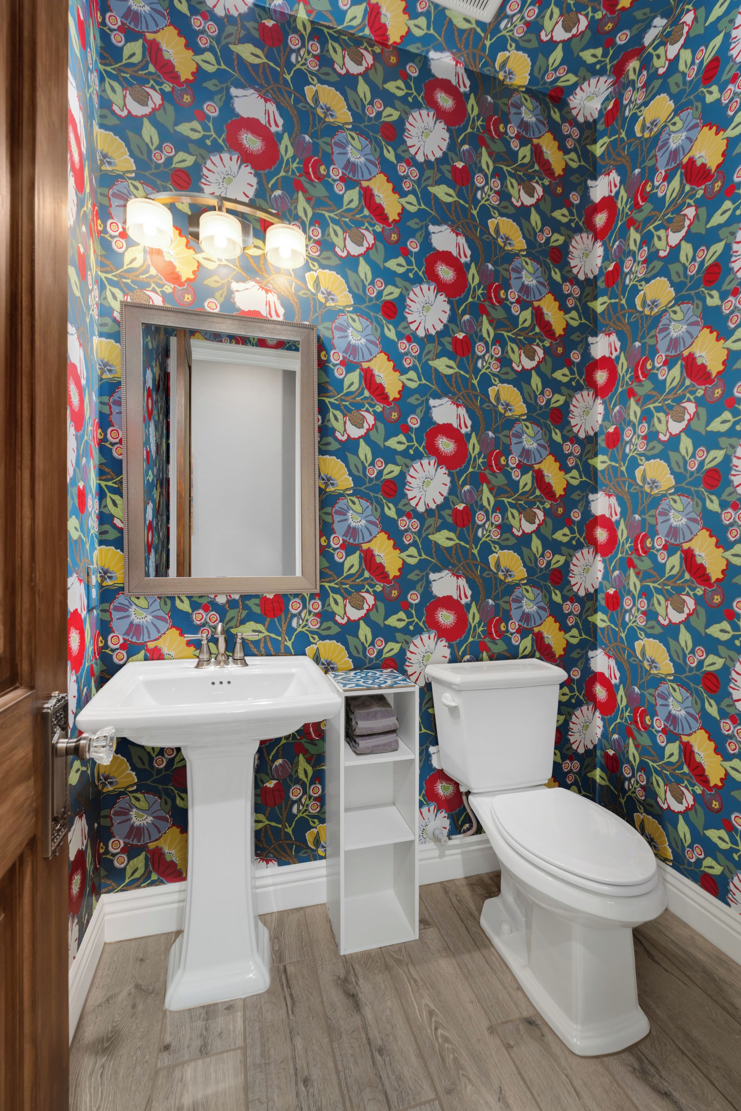 Een badkamer met bloemenbehang, een wit wastafelmeubel, een spiegel en een toilet.