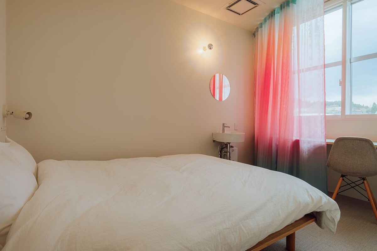 Una cama individual con estructura de madera, una mesita de noche con lámpara, un espejo redondo en la pared, una ventana con cortinas traslúcidas y una silla.