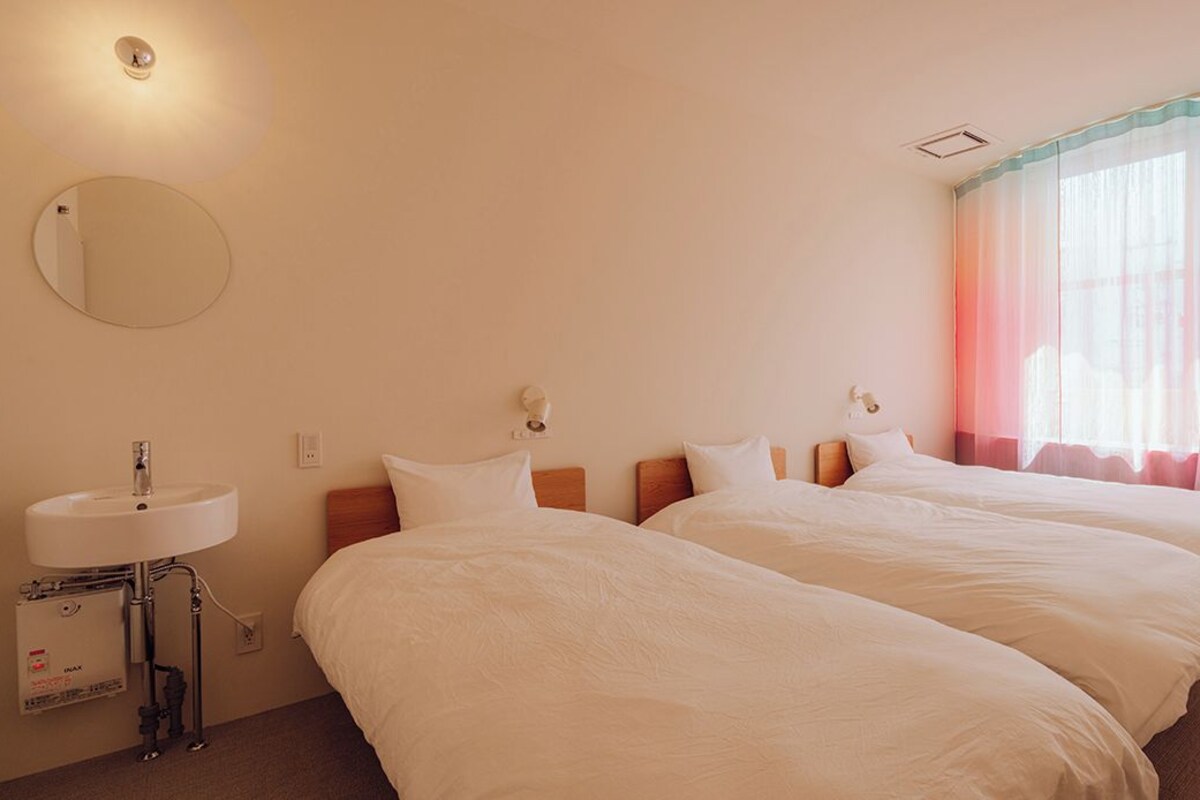 A hotel room with two beds, a round mirror, a sink, and a window with sheer curtains.