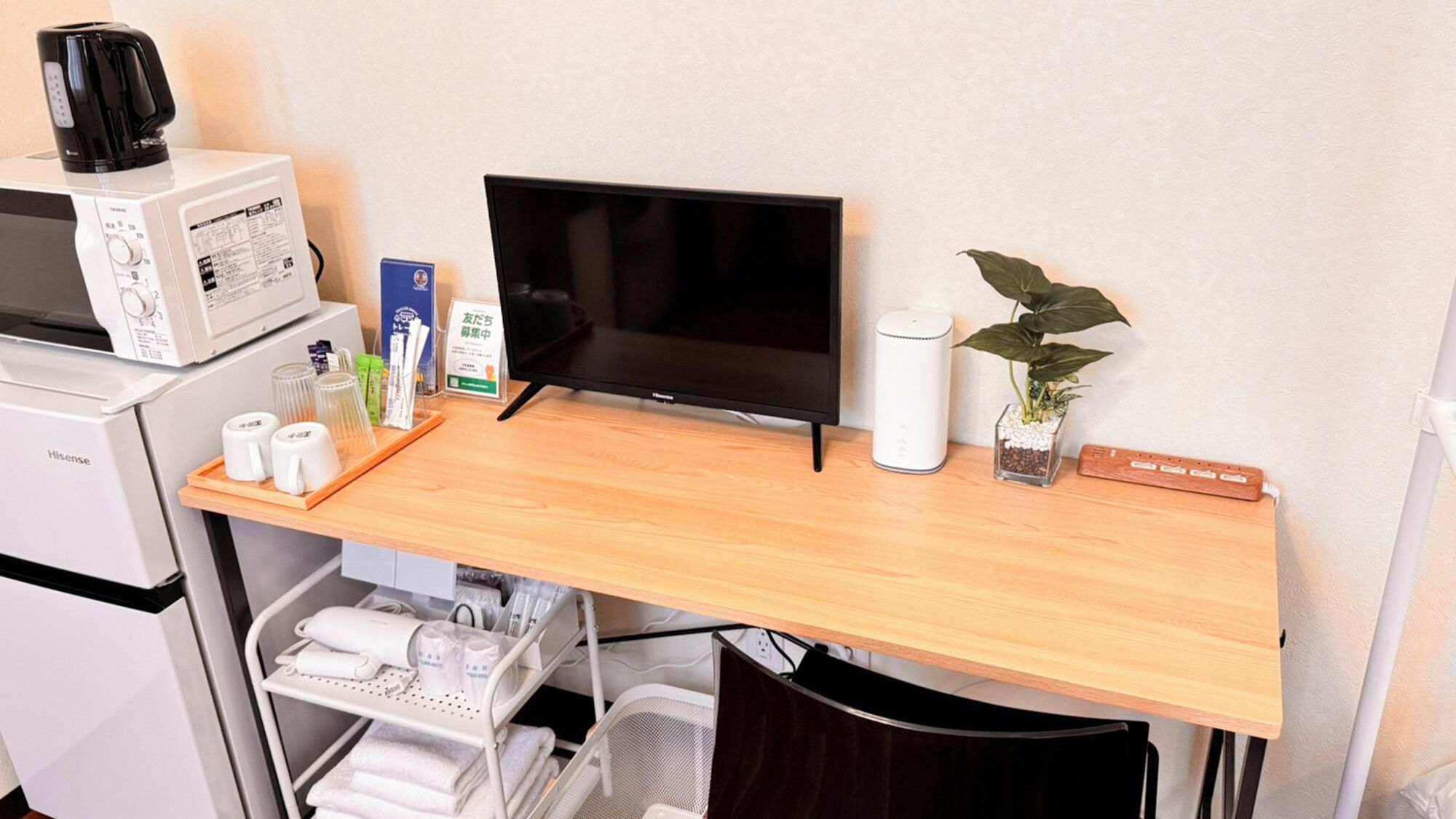 A hotel room with a wooden desk, a flat-screen TV, a microwave, a coffee maker, a plant, and a small cabinet.