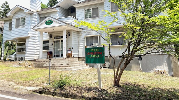 A two-story building with a porch, a green signboard, and a tree in front.