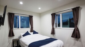 A bedroom with a bed, a nightstand, a TV, and two windows with curtains. - Code Rooms Okinawa (Nakijin)