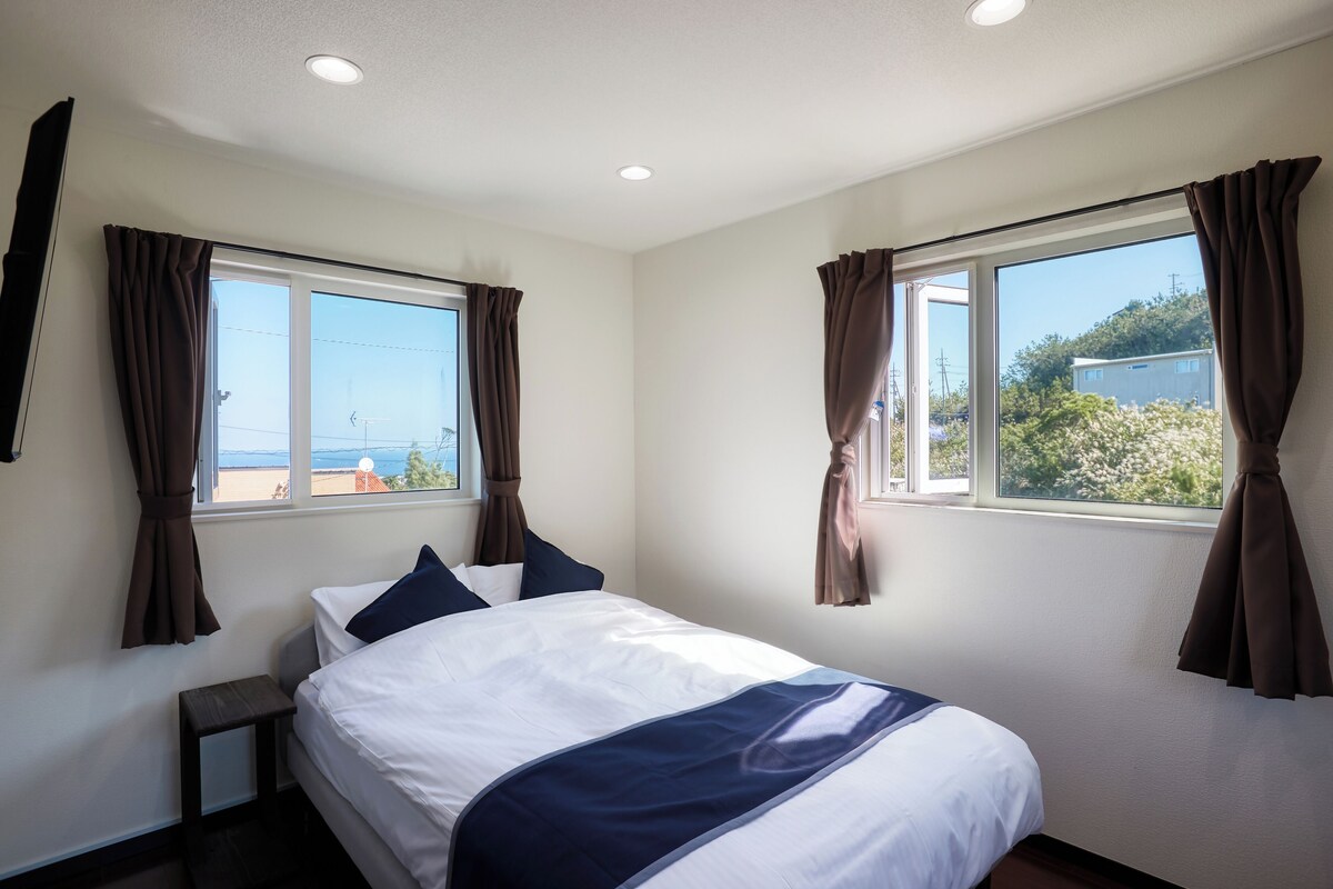 A hotel room with a bed, two windows with curtains, a small bedside table, and a view of the outside.