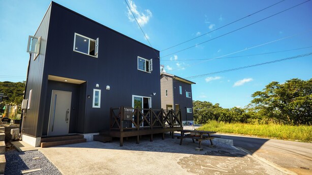 A modern two-story building with a dark facade, a covered outdoor seating area, and a clear sky. - Code Rooms Okinawa (Nakijin)