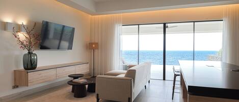 A modern living room with a sofa, a coffee table, a TV mounted on the wall, and a large window with a view of the sea.