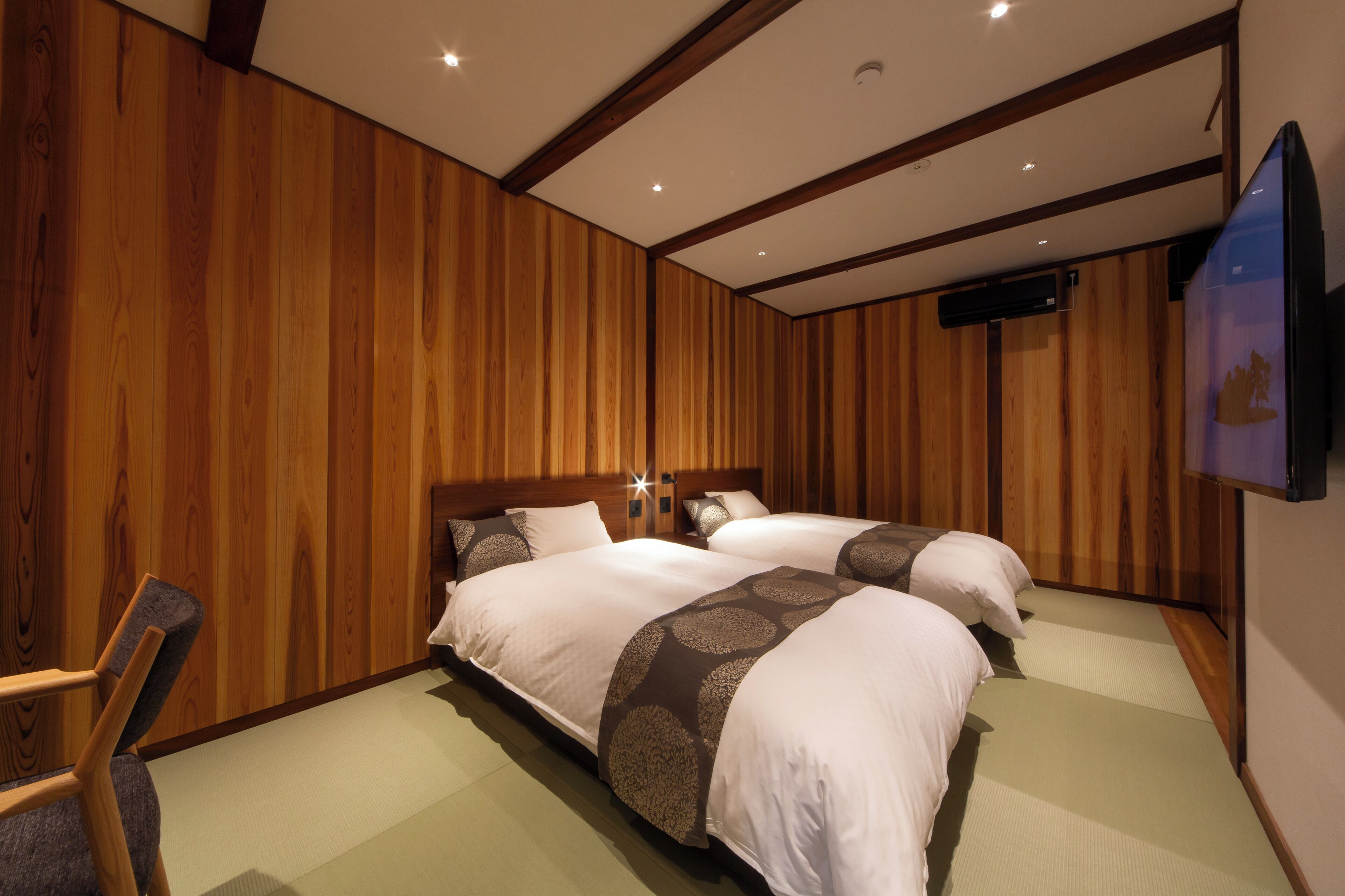 A hotel room with two beds, wooden paneling, a flat-screen TV, and a chair.