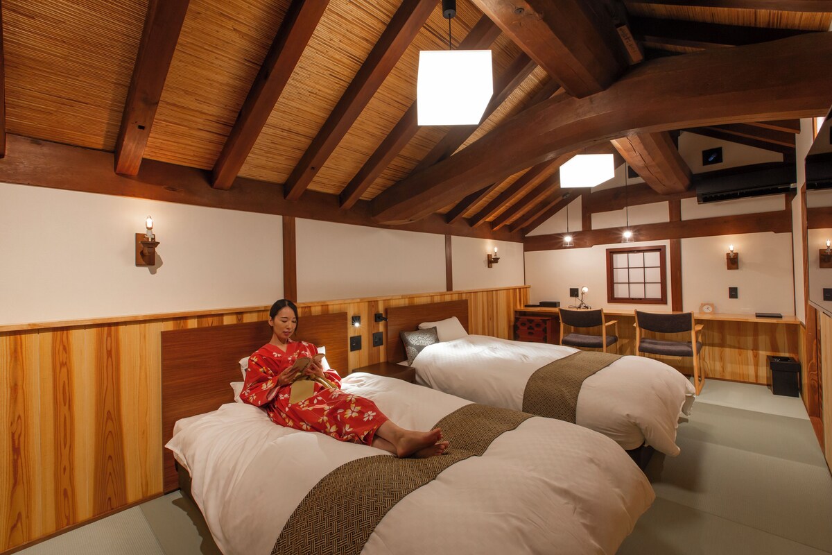 A traditional Japanese-style room with two beds, wooden beams, tatami flooring, and a small desk with a chair.