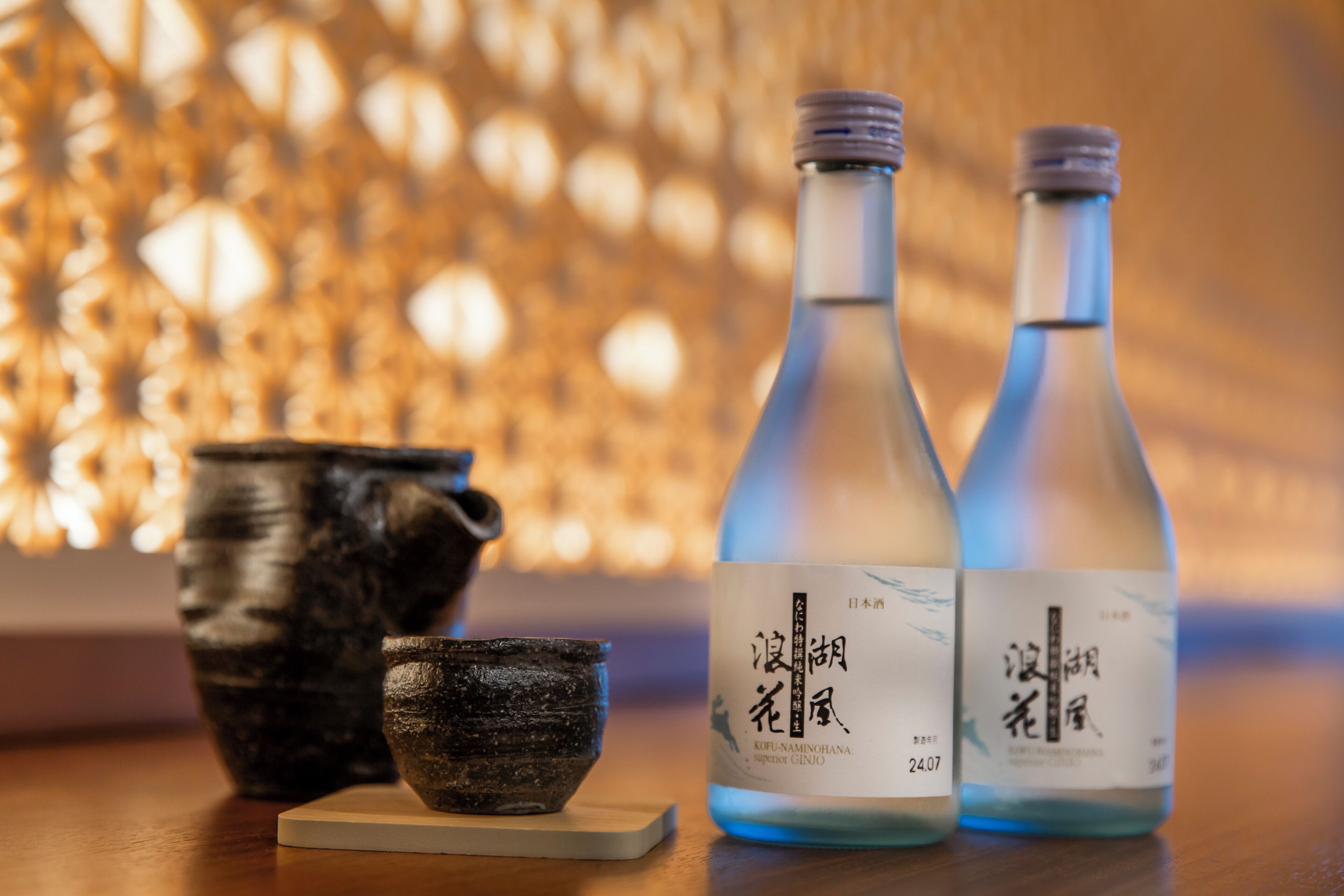 Zwei Flaschen Sake mit Etiketten, eine Keramiktasse und eine kleine Schale auf einer Holztischfläche.