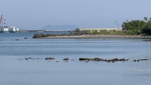 A calm body of water with a distant industrial facility and a rocky shoreline. - Kurinoyakata (Kunisaki)