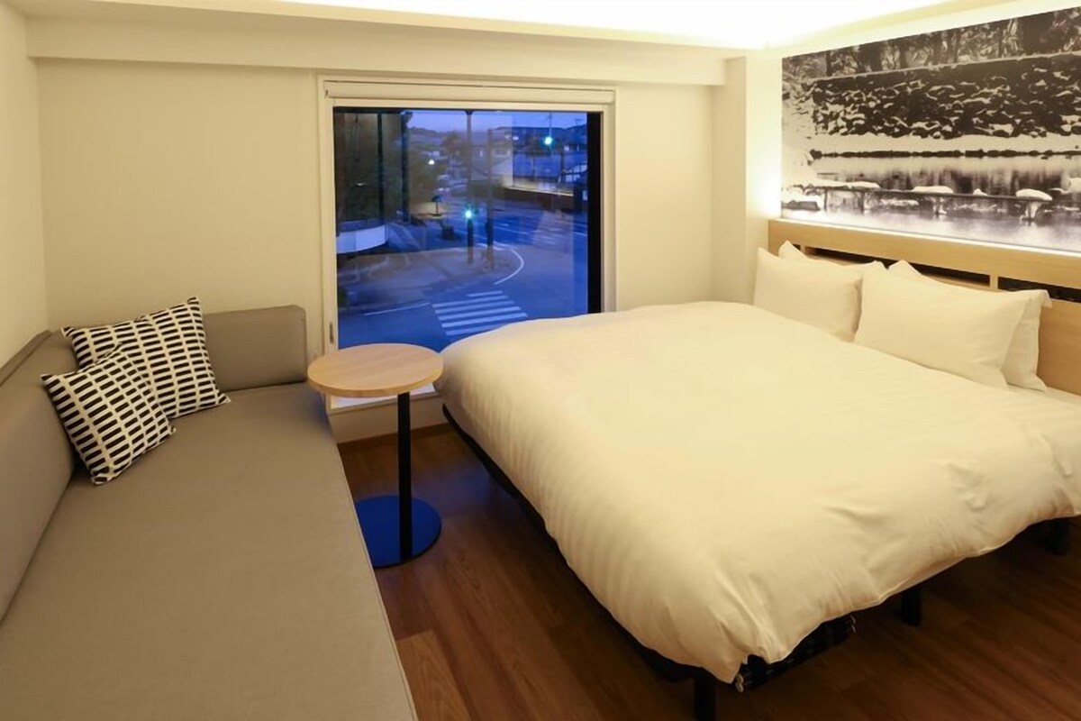 Habitación de hotel con cama, sofá, una mesita redonda y ventana con vista a la calle.