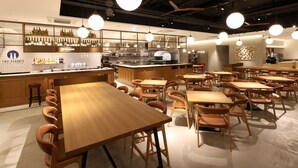 Um café moderno com mesas e cadeiras de madeira, um espaço de bar com garrafas, e uma cozinha ao fundo.