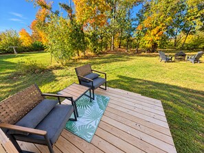 A wooden deck with outdoor furniture, including a sofa and chairs, surrounded by a grassy yard and trees with autumn foliage. - New Build Home near Treetops Golf, Lakes & Trails (Gaylord)