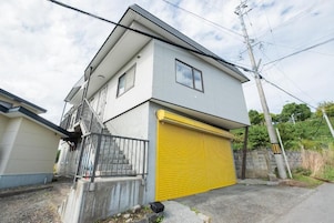Un edificio residencial de dos pisos con una puerta de garaje amarilla, una escalera visible y un muro de hormigón con un pequeño jardín.