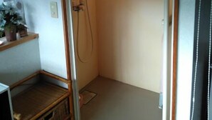 Un box de ducha pequeño con ducha de mano, un estante con una planta y una puerta con panel de vidrio.