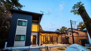A modern two-story building with a visible entrance, a patio area with outdoor seating, and a tent set up in the foreground. - Troll Forest Stella (Nikko)