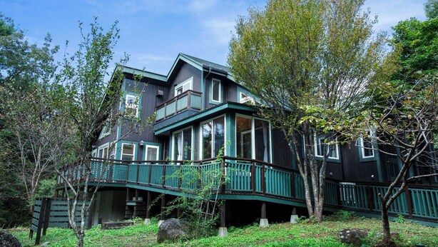 A large, dark-colored house with a balcony, surrounded by trees and greenery.