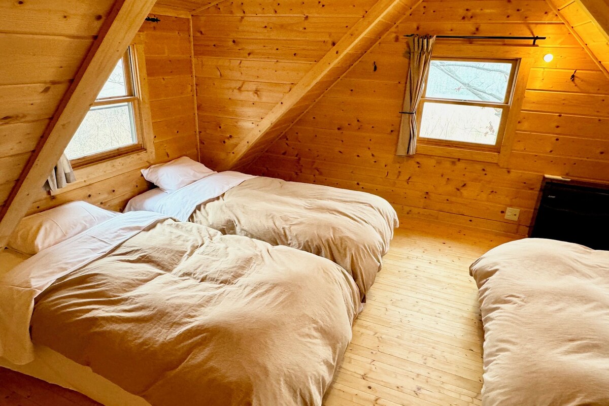 A room with a sloped ceiling, wooden walls, two beds with white bedding, and two windows with curtains.