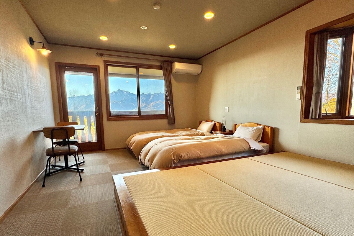 A hotel room with a bed, a desk, and a chair. There are two windows with curtains, and a view of mountains is visible through the windows.