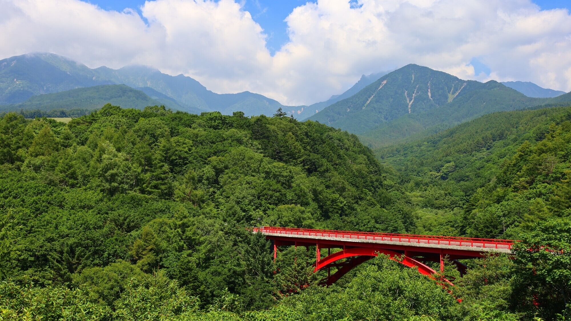 Eine rote Brücke über ein üppiges, grünes Tal mit Bergen im Hintergrund.