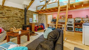 A cozy living room with a stone wall, wooden beams, a fireplace, and a kitchen area with wooden shelves and appliances. - 1 Bed in Llandysul (oc-85671) (Capel Dewi)