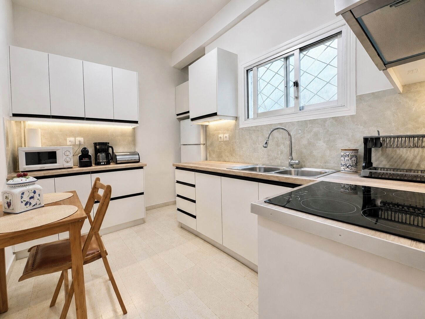 Une cuisine moderne avec des meubles blancs, une plaque de cuisson à induction noire, un évier en acier inoxydable et une fenêtre ornée de vitraux à motifs géométriques.