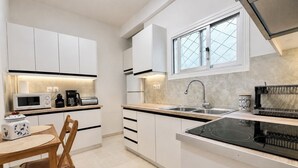 A modern kitchen with white cabinets, a black induction cooktop, a stainless steel sink, and a window with a diamond-patterned glass. - Acropolis Panorama 2BRM Apartment by MΡS (Athina)