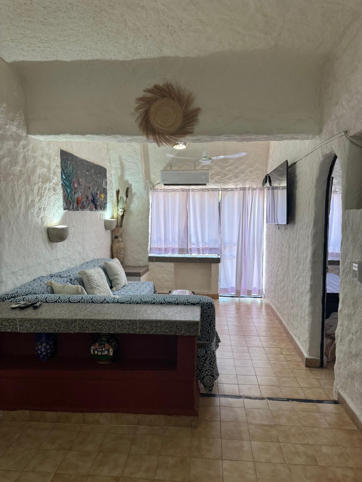 A bedroom with a bed, a painting, an air conditioning unit, a window with curtains, and a television.