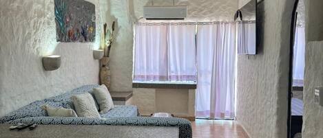 A bedroom with a bed, a painting, an air conditioning unit, a window with curtains, and a television.