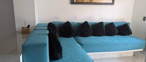A teal sectional sofa with black cushions, a framed artwork above, and a small wicker basket with a phone on the floor.