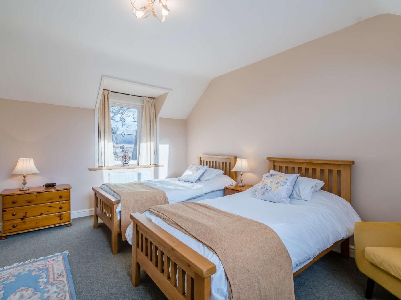 Ein Schlafzimmer mit zwei hölzernen Betten, einem Fenster mit Vorhängen, einem Nachttisch mit Lampe und einem Sessel.