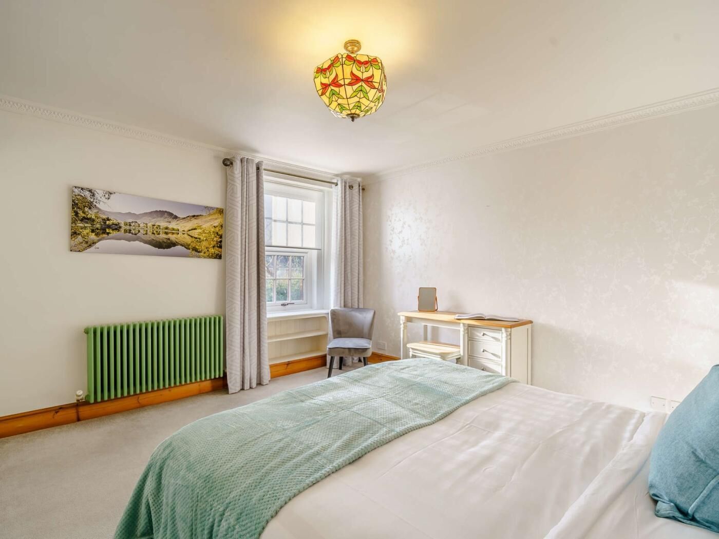 Een slaapkamer met een bed, een bureau, een stoel, een radiator, een raam met gordijnen en een wandlamp.