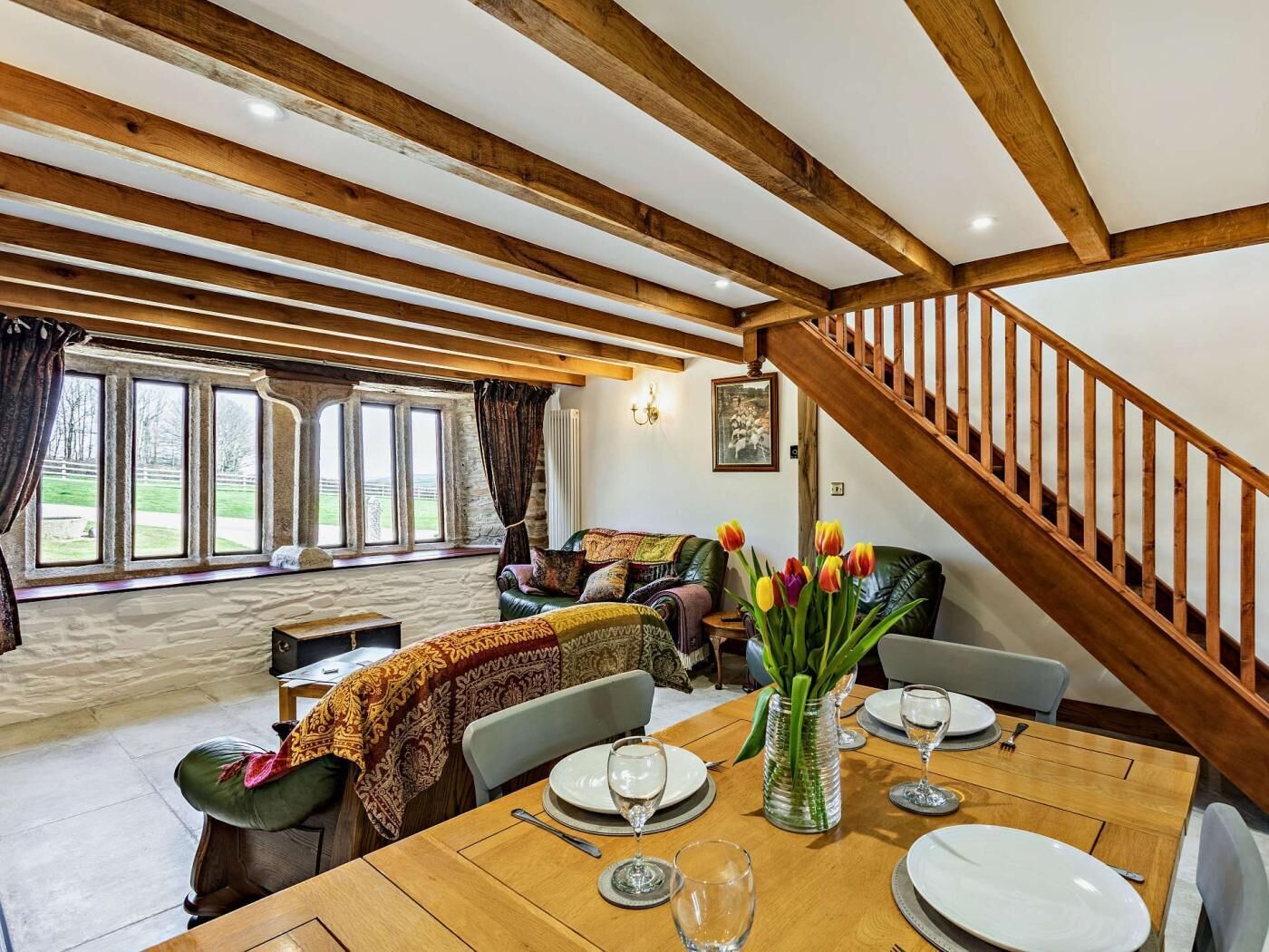 A dining area with a wooden table set for four, a staircase, and a view of a stone wall and greenery outside.