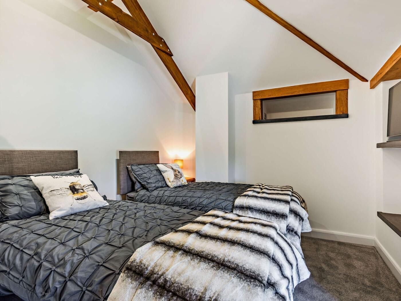 A bedroom with two beds, a wooden ceiling beam, a wall-mounted TV, and a bedside table with a lamp.