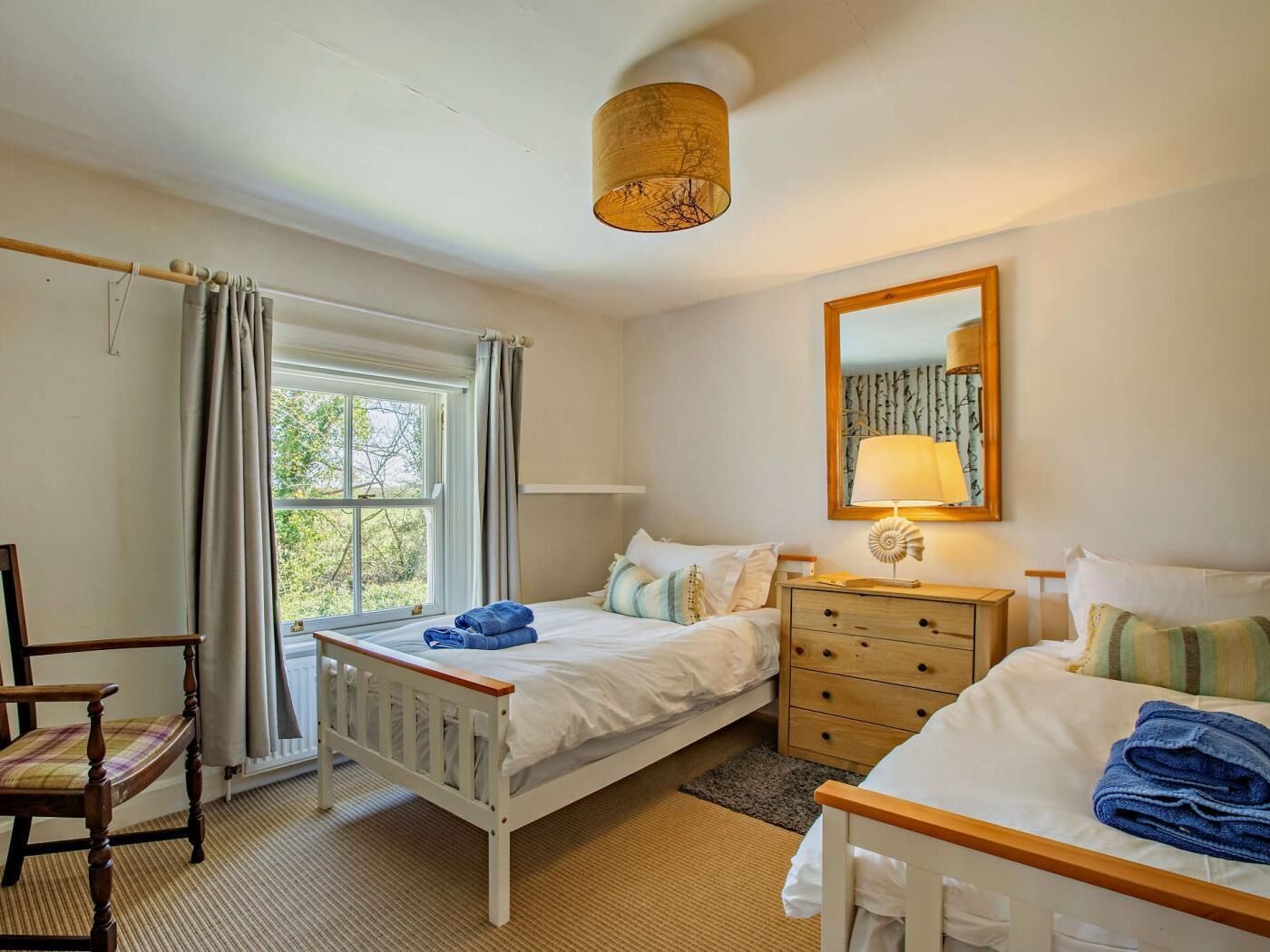 A bedroom with two beds, a wooden dresser, a chair, a window with curtains, a hanging lamp, and a mirror on the wall.