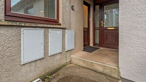 Ein Gebäudeeingang mit einer Holztür, einem Fenster mit Rollläden und drei Stromkästen an der Wand.