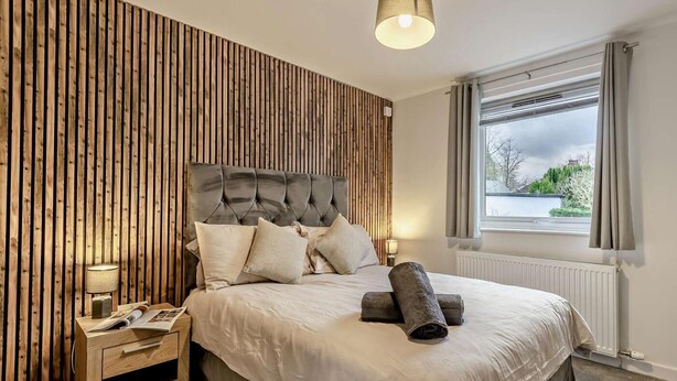 Ein Schlafzimmer mit einer Wand aus Holzlamellen, einem gepolsterten Kopfteil, einem Bett mit weißen Bettwäsche, einem Nachttisch mit einer Lampe und einem Fenster mit Vorhängen.