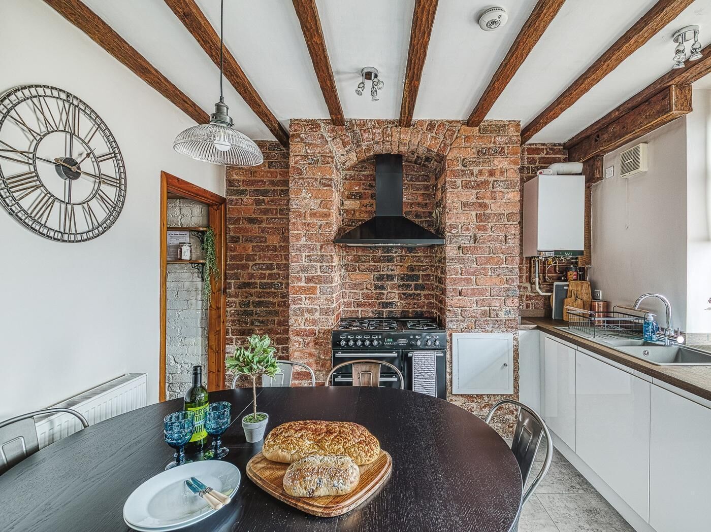 Una cocina con pared de ladrillo, vigas de madera, un reloj y una mesa de comedor con pan.