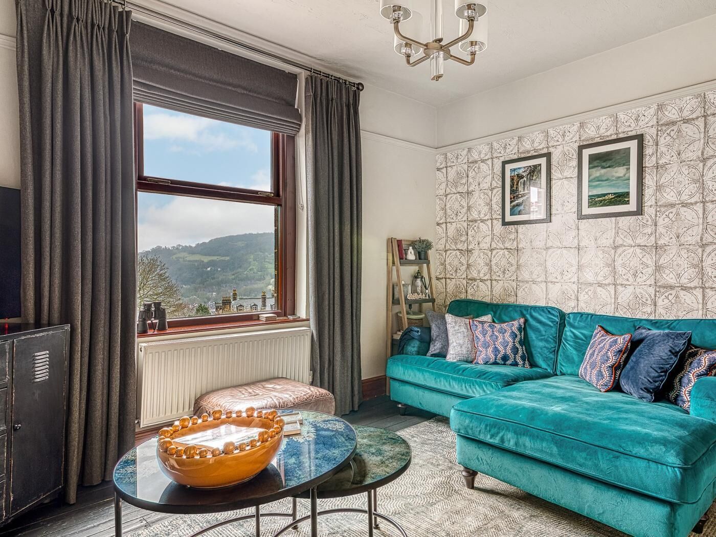 A room with a teal sofa, a wooden sideboard, a large window with a view, and patterned wallpaper.