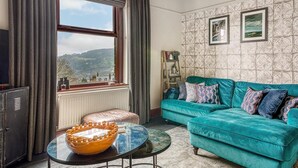Ein Zimmer mit einem petrolfarbenen Sofa, einer Holzkommode, einem großen Fenster mit Blick und Wandpapier mit Muster.
