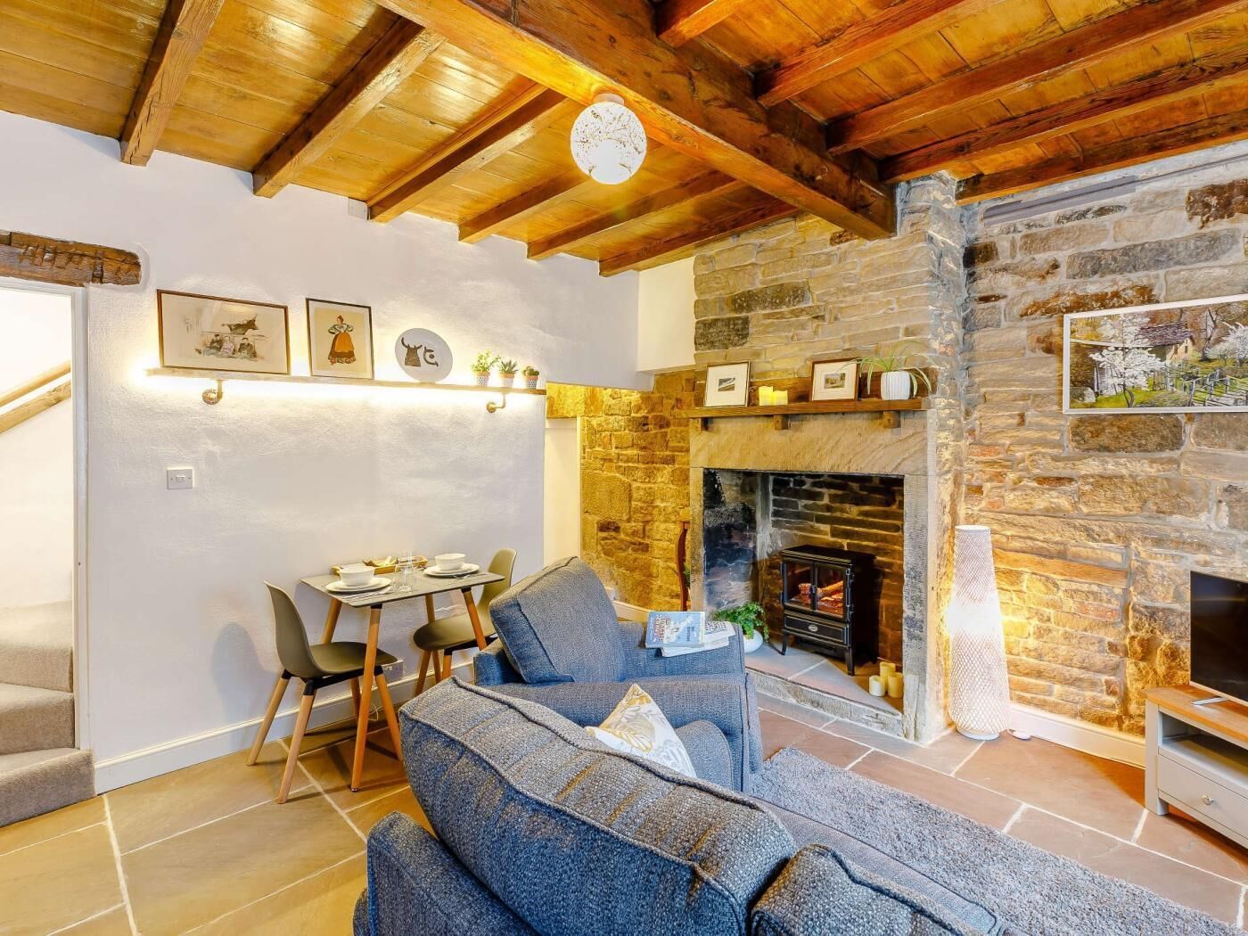 A cozy living room with a stone fireplace, wooden ceiling beams, and a dining area with chairs and a table.