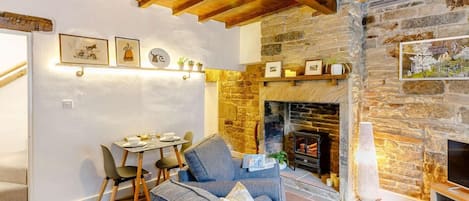 A cozy living room with a stone fireplace, wooden ceiling beams, and a dining area with chairs and a table.