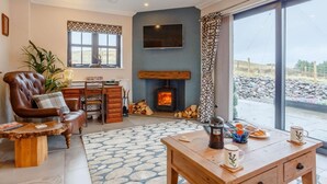 A cozy living room with a fireplace, a comfortable leather chair, a wooden coffee table, and a large window offering a view of the outdoors.